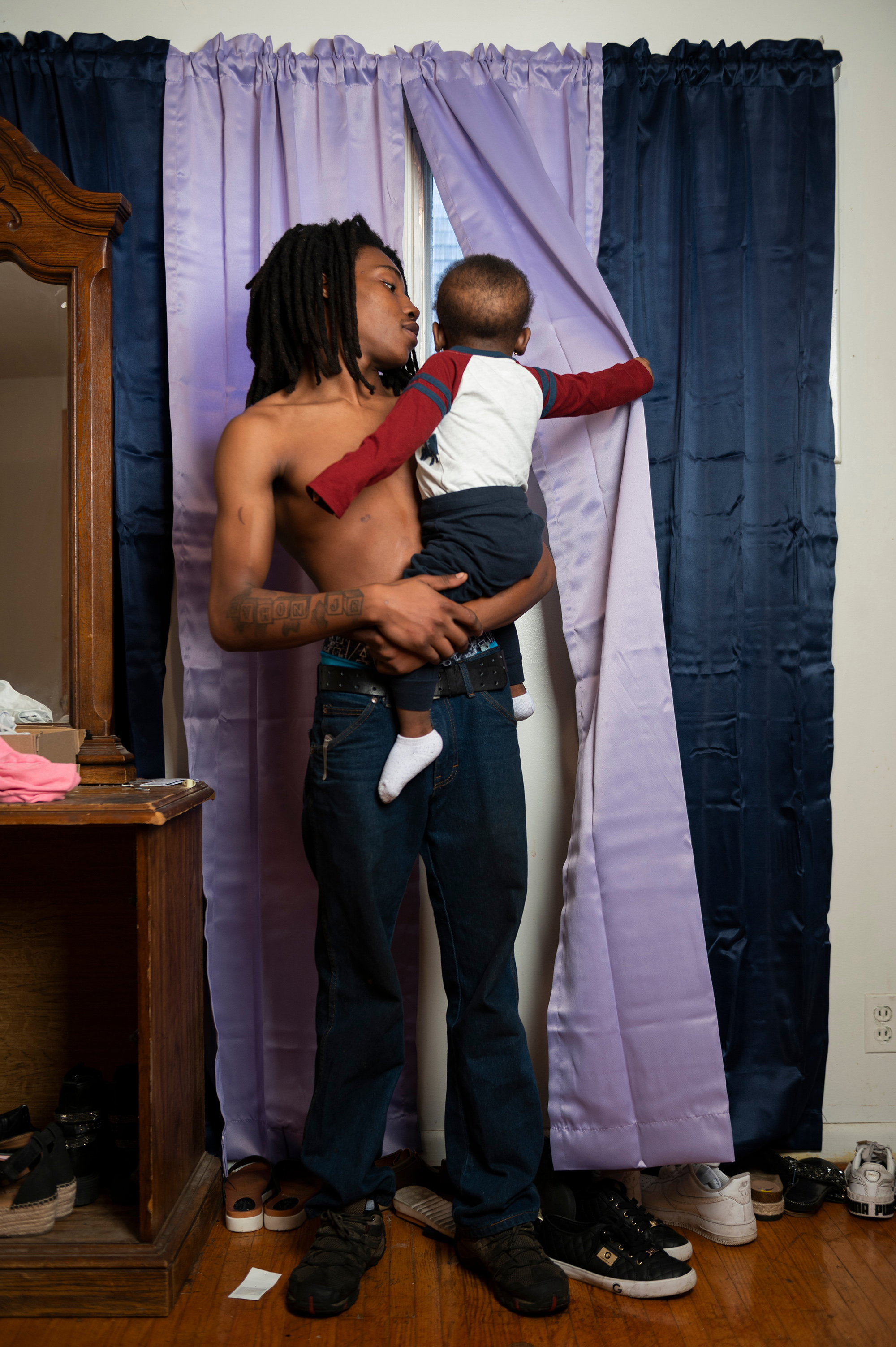 A man holds a little boy in his arms as the boys peels back a curtain to look out the window.