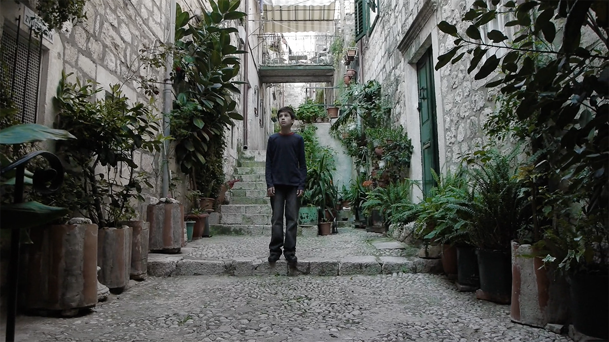 A film still of a young boy in an alley surrounded by plants.