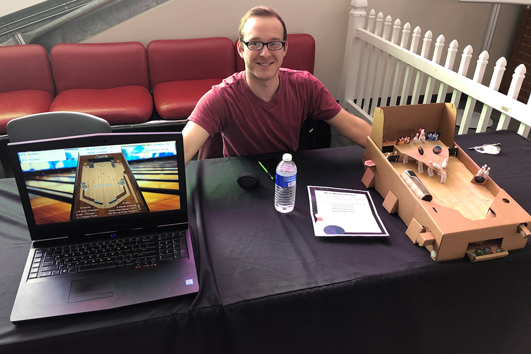 student sitting at table with cardboard prototype of pinball machine and laptop displaying virtual pinball machine.