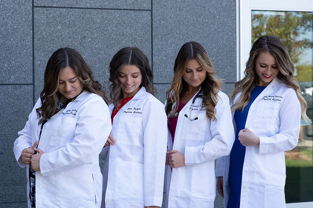 four students look at the names embroidered on their white lab coats.