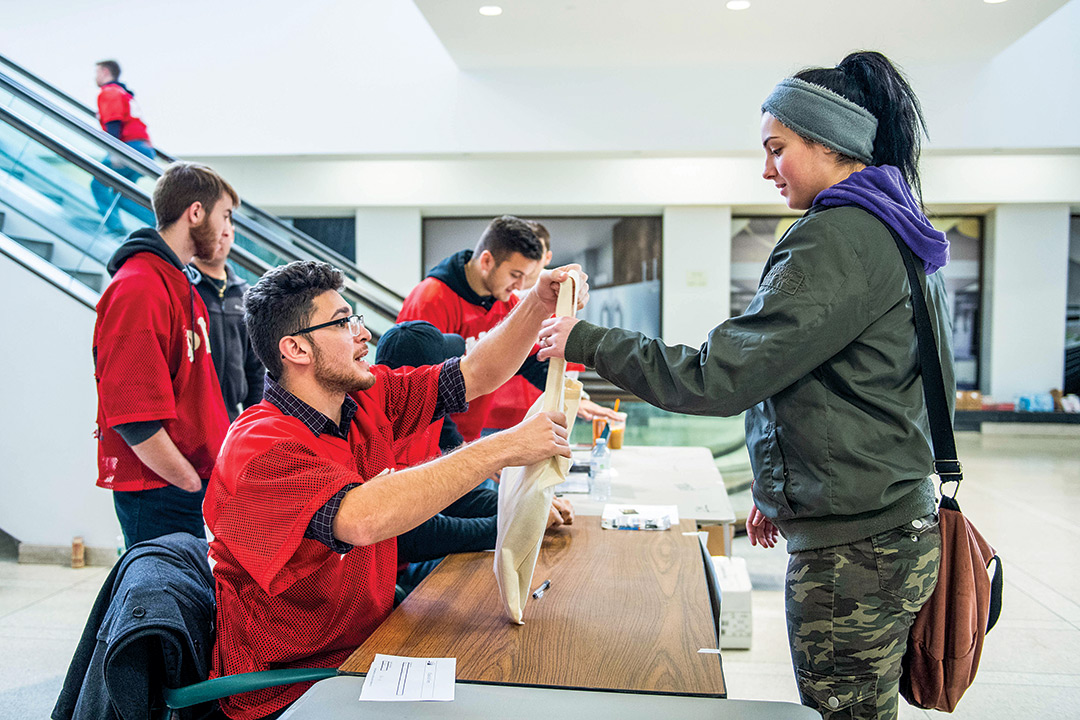 Student in red shirt hands bag to woman standing at table.