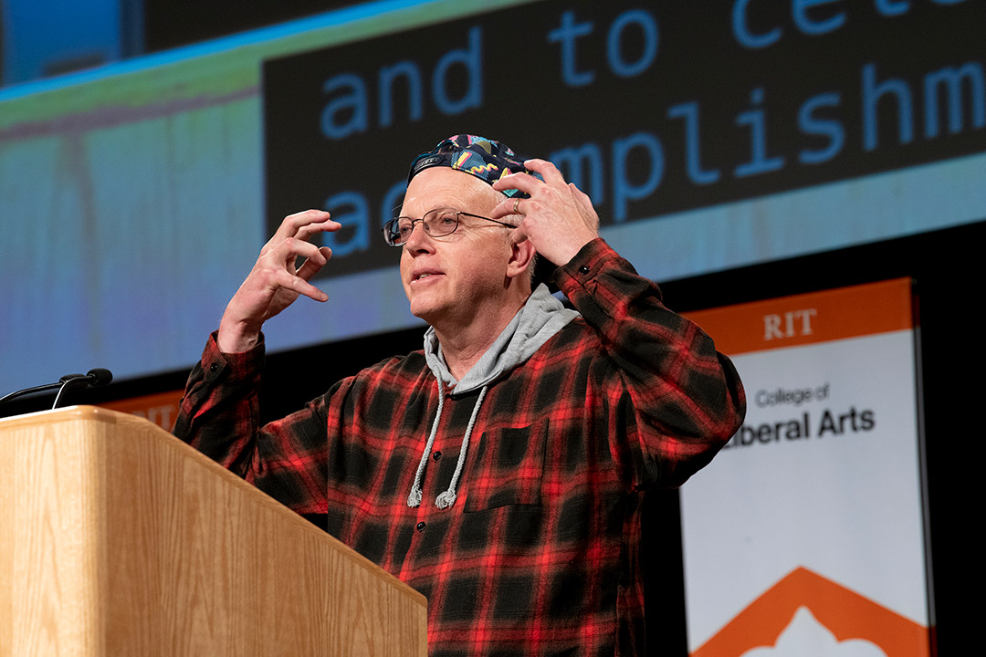 RIT President Munson on stage wearing a backwards baseball hat and a plaid hoodie.