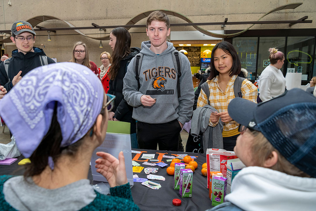 two students standing talking to two seated students at an information table.