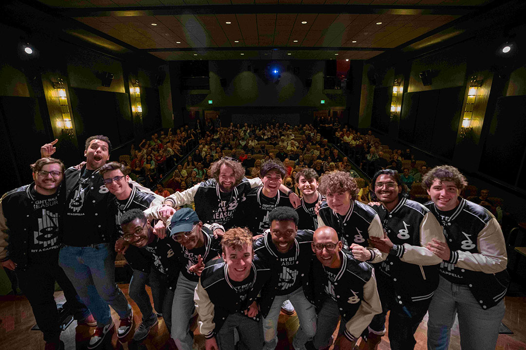 group of college students wearing varsity jackets posing for a photo on a stage with a crowd seated behind them.