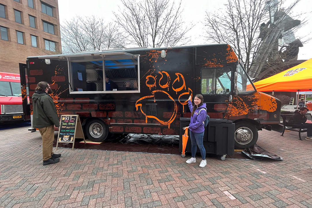 two college student standing outside of a food truck painted black with orange splashes and a brick design.
