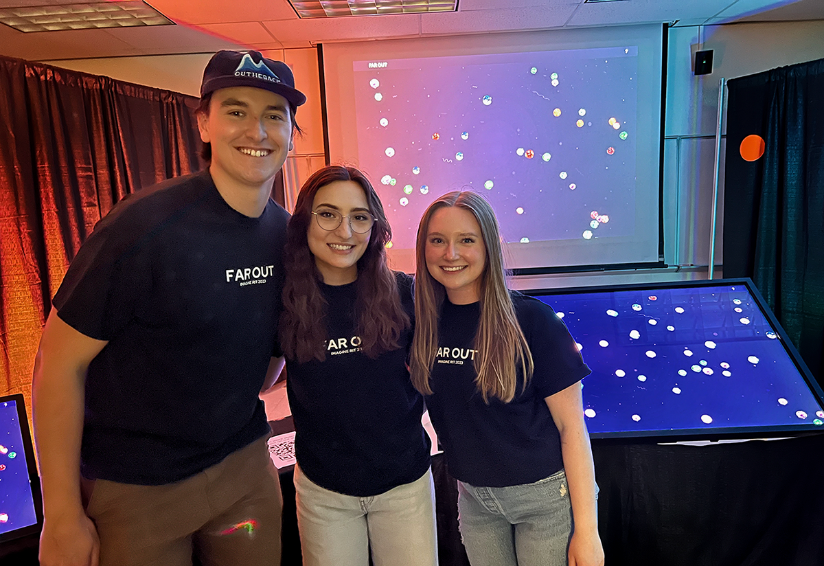 A group photo of three students standing in front of touchscreen displays.