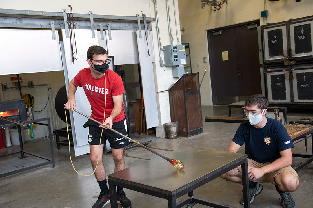 student observing another student blowing glass.