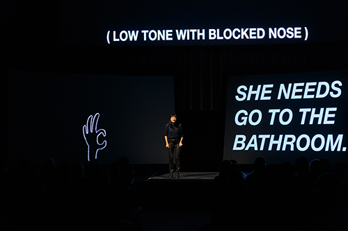 Kim, wearing black clothes, stands on a stage, dwarfed by three large screens surrounding her. To the left, a black screen displays a hand-drawn hand with thumb and index finger forming a circle, drawn in white; above her is, in white, text reading "(Low tone with blocked nose)"; on the right side of the image is the third screen, which reads in large white text "She needs to go to the bathroom."