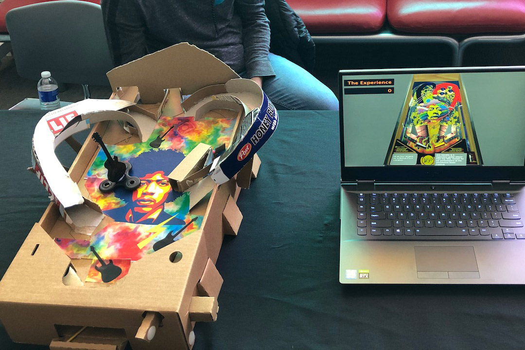 cardboard prototype of pinball machine next to laptop display of pinball machine.