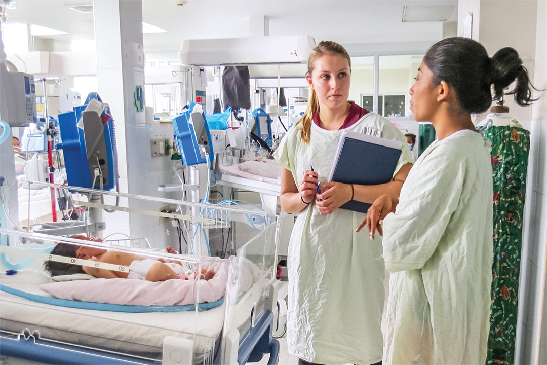 Two women in scrubs stand talking next to isolette with baby.