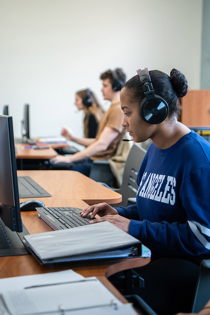 Student sits at desk with computer and headphones.