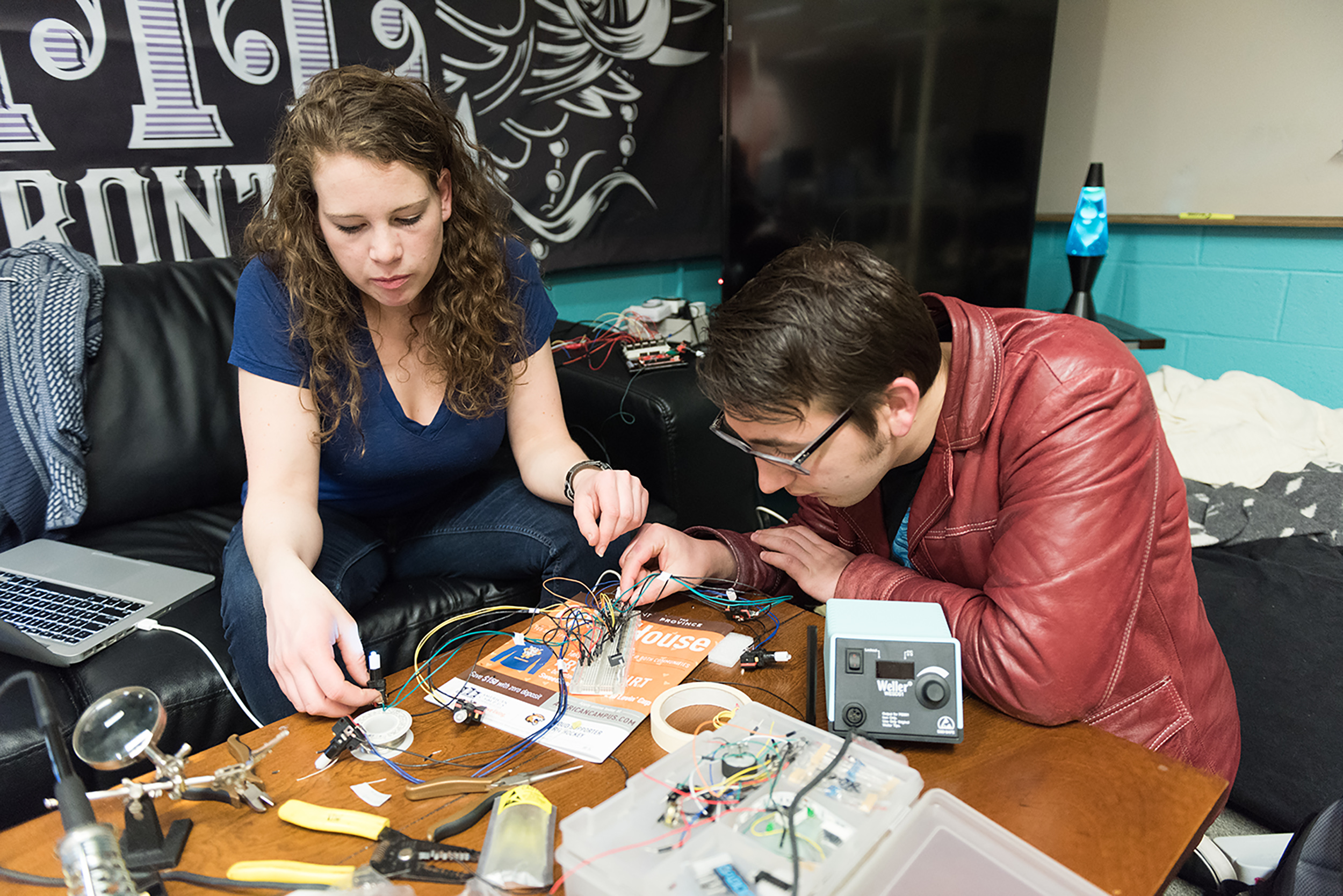 Two students sit at a table with a variety of electronics while soldering some of them