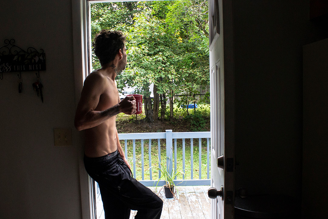 man wearing sweatpants holding a mug of coffee learning against a doorframe looking outside.