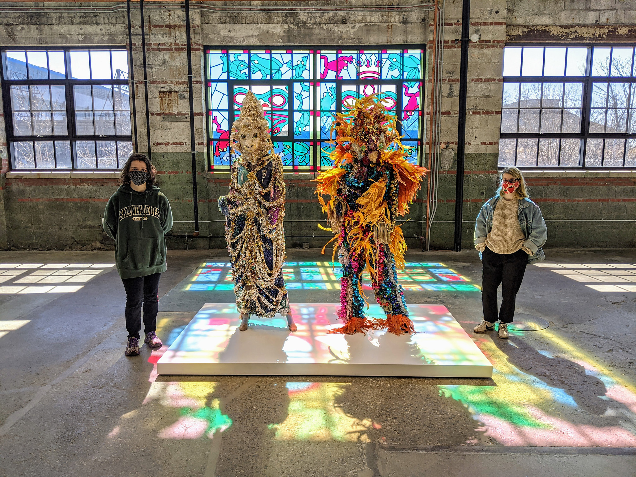Two students stand beside two large sculptures inside a warehouse-looking space.