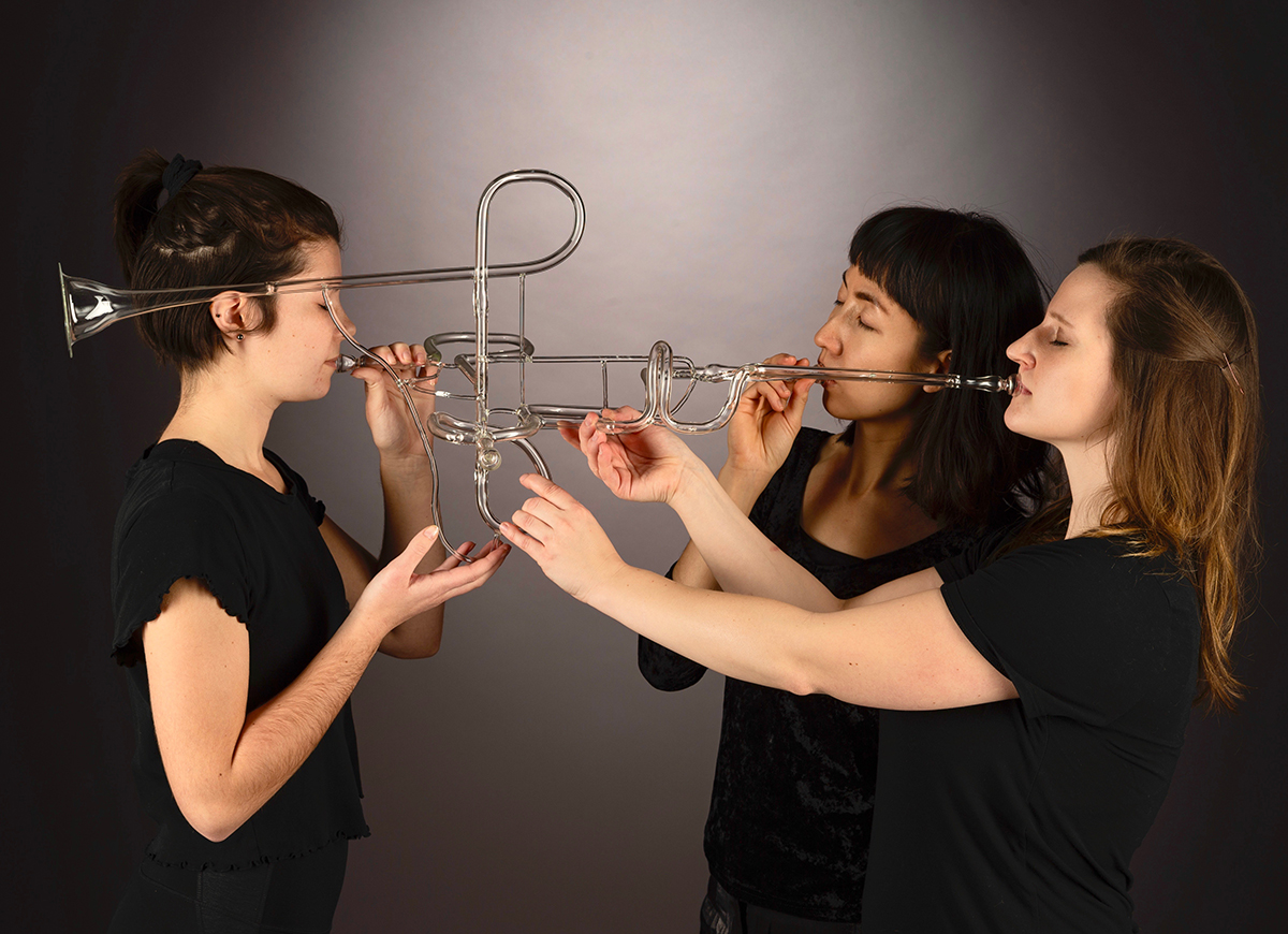 Three students blow into mouthpieces of a flameworked trumpet.