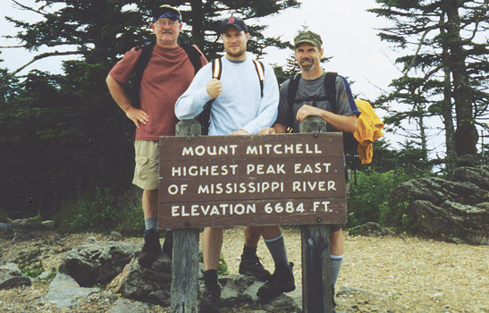 Three people standing behind sign "Mount Mitchell Highest peak East of Mississippi River Elevation 6684 ft"