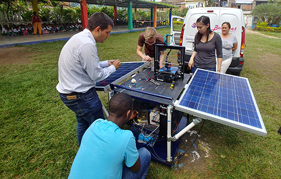RIT students and Professor Rojas are outside setting up their solar powered 3D printer. In the distance, a large group of school children look at their progress.