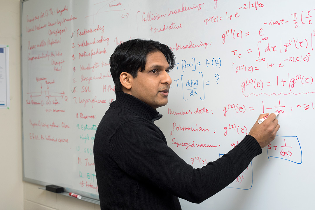 Mishkat Bhattacharya standing in front of white board and writing out equations.