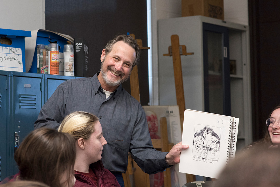 Leigh Rubin holds up a sketch and smiles at the camera as students look on.