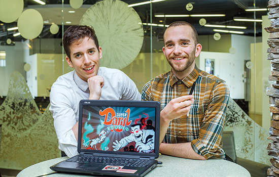 2 students sitting at a desk, showcasing their game on a laptop screen.