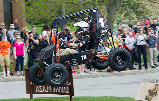 A student drives the Baja Racing vehicle as the crowd looks on. The car sails through the air after driving over a ramp.