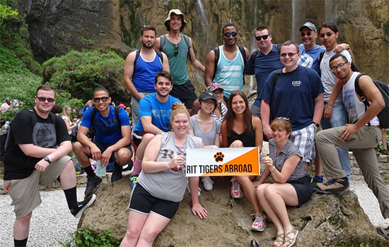 A group of students who traveled abroad pose for a photo on a large boulder holding up a poster that reads "RIT Tigers Abroad."