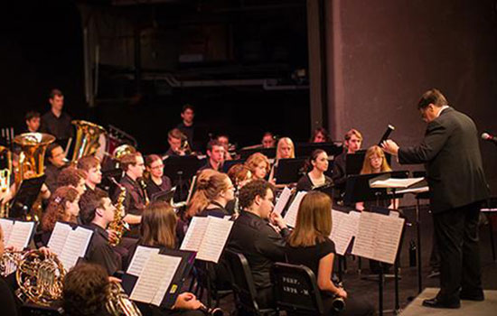 A photo of RIT's concert band performing. The conductor stands on a short platform at the front and uses his hands to instruct the players. 