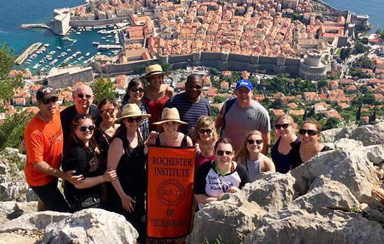A group of RIT students and parents pose on the mountainside in Croatia, a beautiful city landscape behind them. They hold a banner that reads "Rochester Institute of Technology."