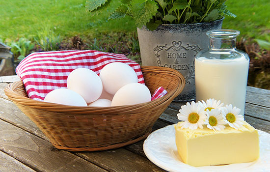 An artistic photo of eggs in a basket, milk in a glass bottle, and a large block of cheese with daisies on top of it.