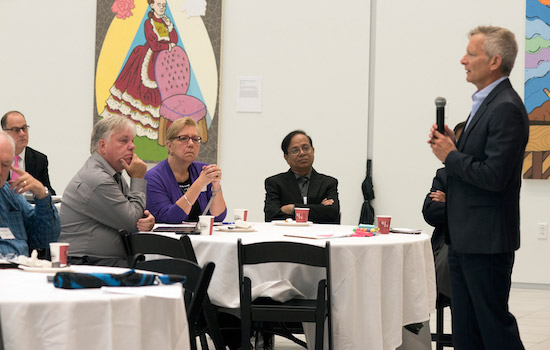 One person stands at the front of the room talking into a microphone while the audience sit at tables and look at him, engaging in what he is saying.