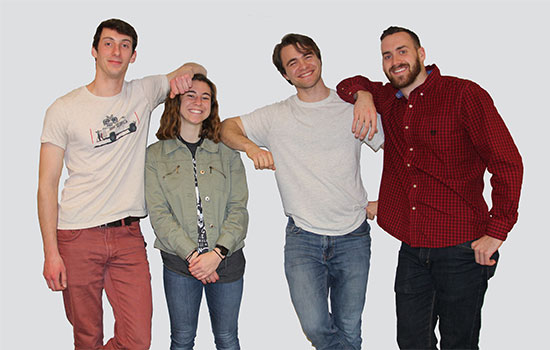 Four members of the Gaia team stand together, posing for a photo by leaning against each other's shoulders with their elbows. James has his elbow on Kerri's head to accentuate their height difference.