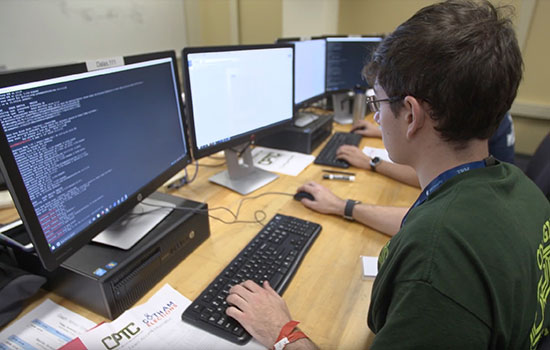 A student sits in front of a computer and writes out code, which is visible on the desktop screen.