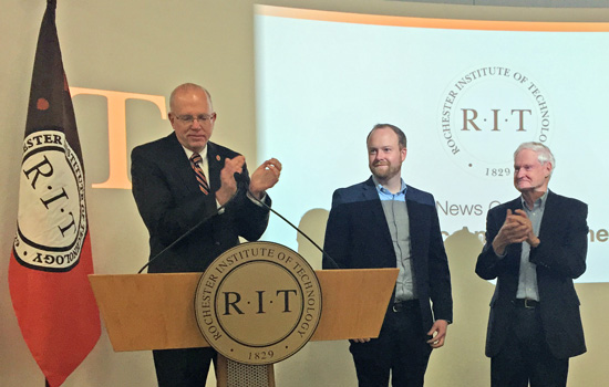 Person clapping standing behind a podium, accompanied by two other people.