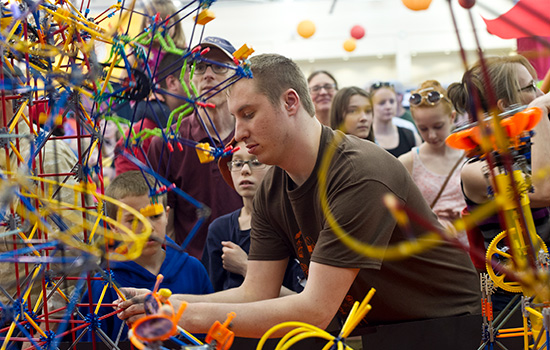 A student at Imagine RIT adjusts the toy-model of a theme park while children and their parents look on behind him.