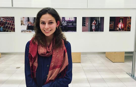 Lauren Peace poses for a photo in the University Gallery. She stands in front of a board with five photos lined up next to each other.