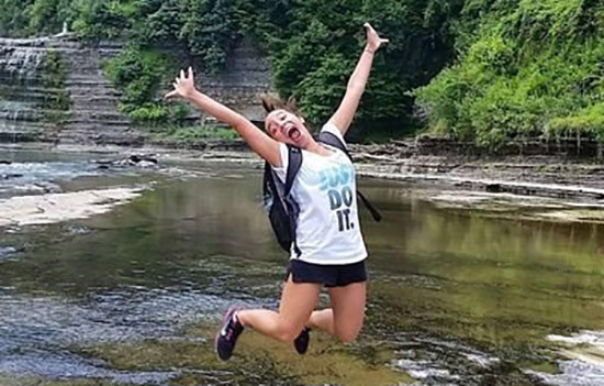 Nina Hullett jumps up in the air, extending her arms and smiling wide. She is outside near a large creek, seemingly on a hike.