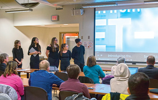 The Jet-2k team stands at the front of a room and shows students and potential "investors" a video about their packaging design.