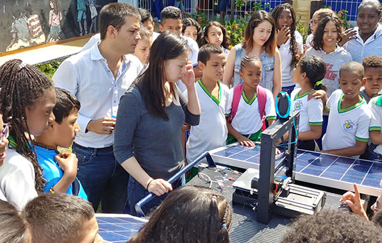 Team members demonstrate their new 3D printer and young students crowd around them to look and see the machine function.