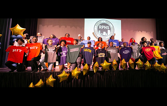 Students on stage holding up their future colleges' shirts.