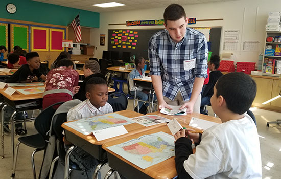 A person talking to two kids in a classroom. Kids are sitting at desks with maps of America in front of them.