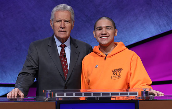 Sheldon Lewis smiles next to Alex Trebek, wearing a bright orange RIT sweatshirt.