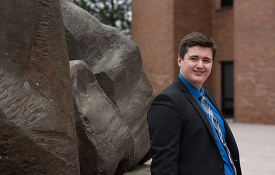 Zachary smiles at the camera, leaning against an outdoor sculpture behind the College of Liberal Arts.