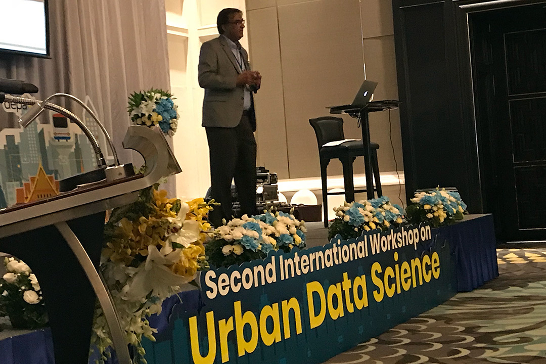 A man stands on stage and talks to a group of people. Attached to the edge of the stage is a banner that reads "Second International Workshop on Urban Data Science." There are also several flower arrangements scattered across the stage.