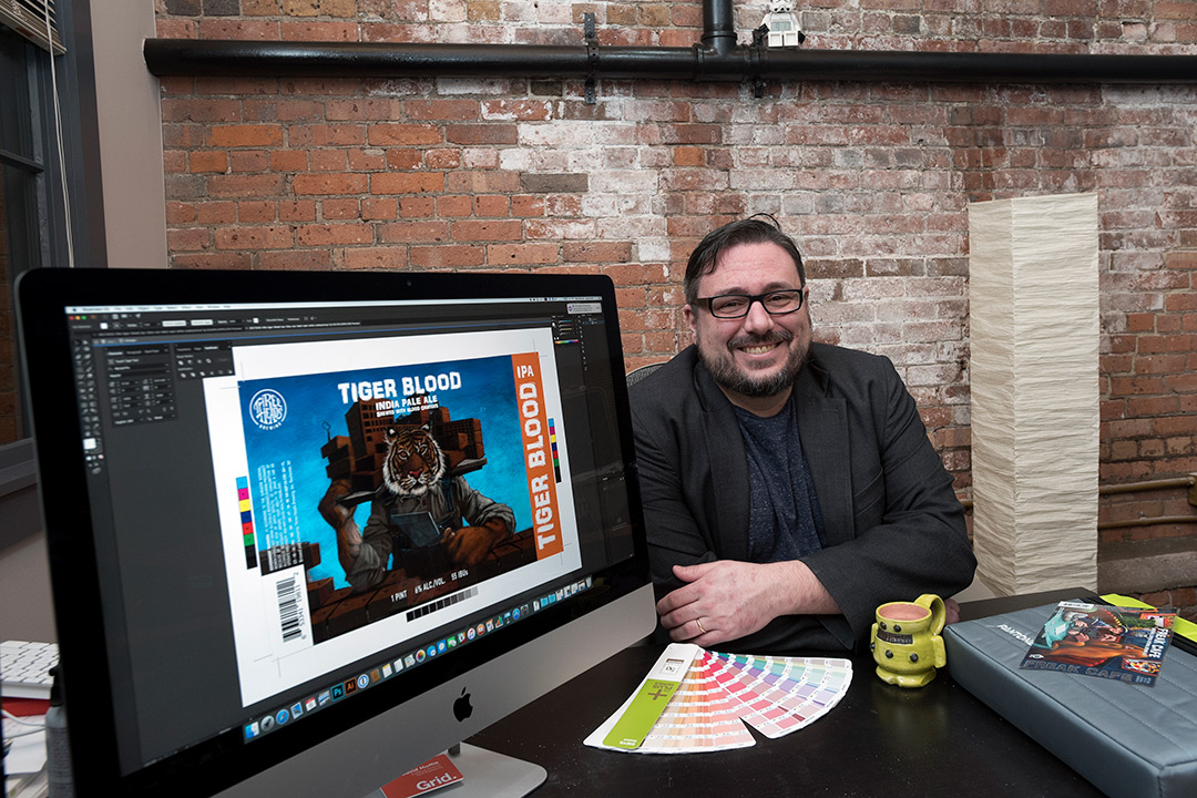 David Moffittt poses for a photo at his office desk. His computer is turned so the screen is facing the camera and the design for the beer label is pulled up on photoshop.