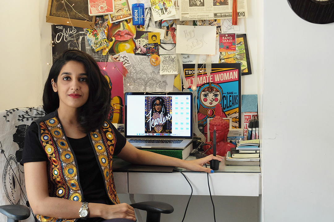 Shehzil Malik sits at her desk and poses for a photo. Her desk and wall are covered in colorful artwork.