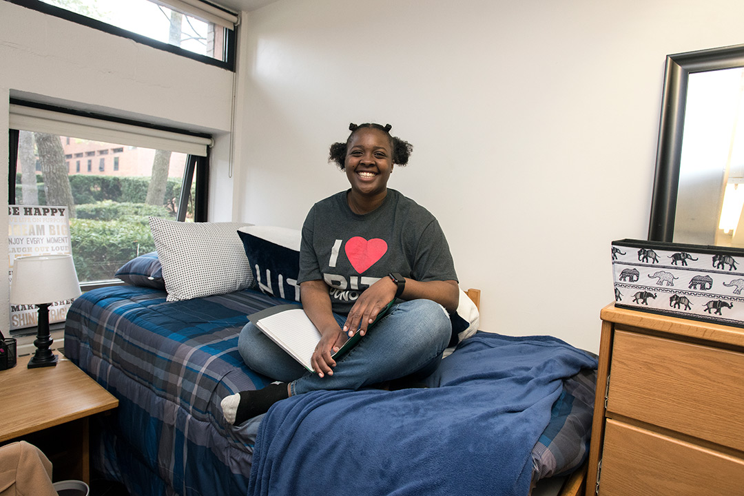 Shantinique Cowans sits on her bed in her dorm room, smiling and holding a book open in her lap.