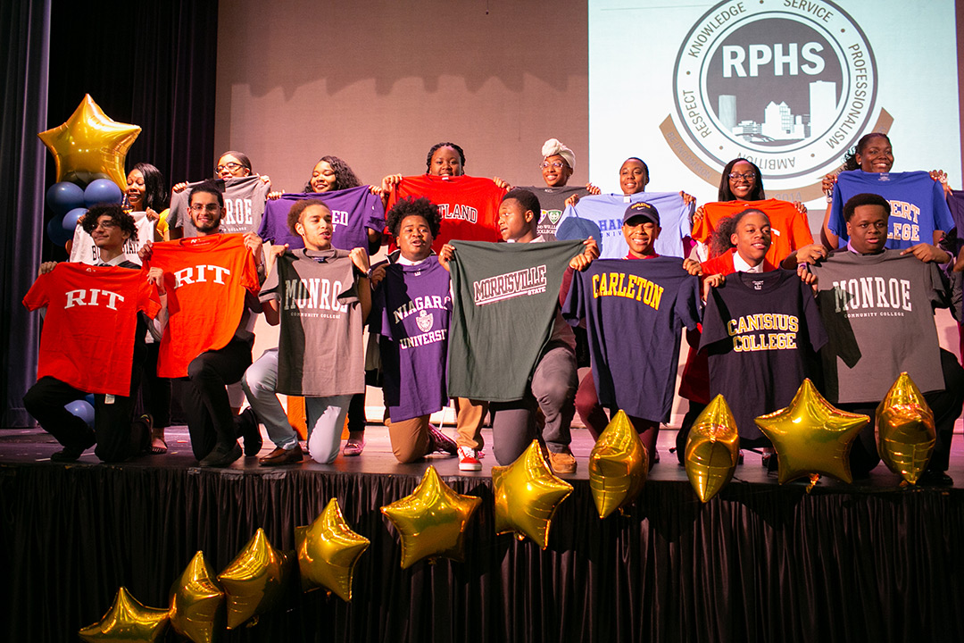 A group of graduating students from Rochester Prep High School stand on a stage and hold up t-shirts from the colleges they have been accepted into for the fall.