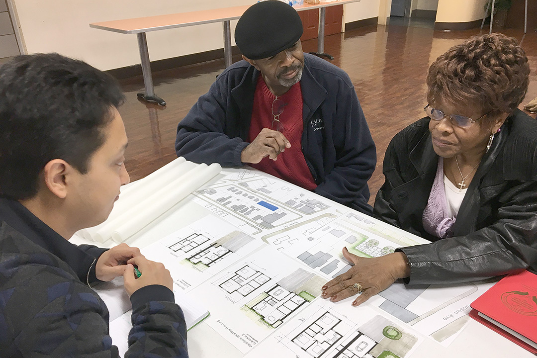 Madi Zhunussov discusses floor plans and designs with two representatives from the Coalition of NorthEast Association. They sit across from each other at a table with the designs covering the table top.