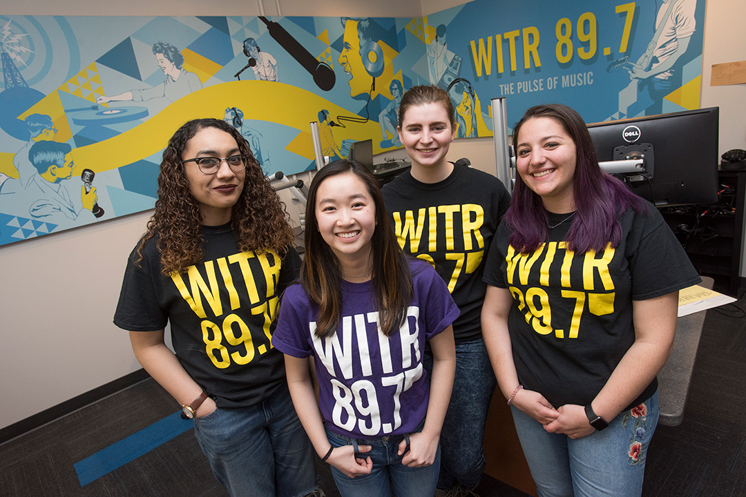 The four women e-board members stand together in the stations broadcasting room.