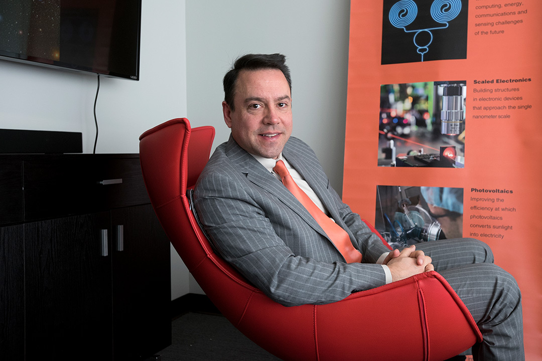 A portrait of Don Figer. He is seated in a contemporary red chair and he is leaning on the arm of the chair.